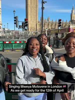 A group of three women stand outside. They are looking into the camera and smiling. The background includes a glimpse of a large building with a clock tower and a bustling street, hinting at an urban setting. The sky visible above is clear and sunny, while a banner across the bottom of the image provides additional context.