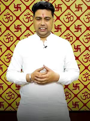 A man in a white shirt is centered in the frame, hands clasped. The background is a repeating pattern of yellow ochre and red-brown symbols reminiscent of an ancient script or decorative motif. His hair is dark, and he has a small red marking on his forehead. The composition is a medium shot, focusing on the upper body of the man.