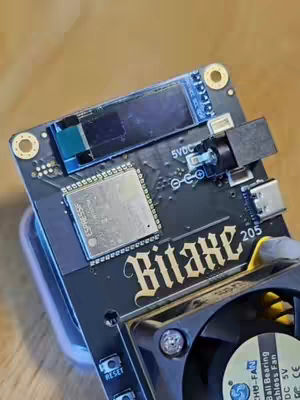 The cover image shows a close-up of a black circuit board with various electronic components. The main focus is on the "Bitaxe" logo in gold lettering. A small fan is visible at the bottom, and the background is a blurred wooden surface. The overall aesthetic is technical and informative.