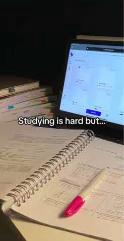 A dimly lit scene dominated by study materials. A spiral notebook and a pen with pink ink are in the foreground, alongside a tablet displaying a webpage. Stacks of books are visible, suggesting an academic setting.