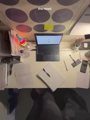 An overhead shot of a desk with a laptop open, papers, pens, and a calculator. The ceiling above has large circular designs.