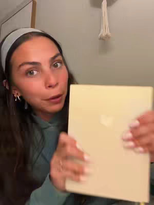 A woman with long dark hair holds up a closed bible towards the camera. She is wearing a light teal hoodie and a headband.