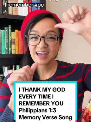 A woman with dark hair and glasses wears a red beret and a striped sweater, smiling and pointing with her right index finger. Text in a blue box below reads a Bible verse and states a memory verse song.