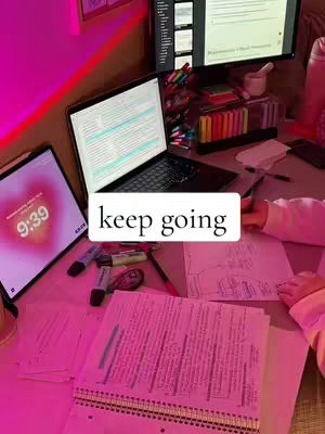 A well-lit workspace is shown with several items on the desk: a laptop, a tablet displaying a heart-shaped clock, and various stationery items. The desk is bathed in a pinkish hue, suggesting ambient lighting. In the foreground, there is an open notebook with handwritten notes. The image has a focus on productivity and study.