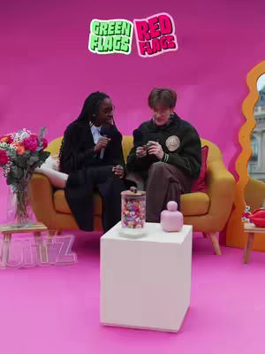 The cover image captures a scene inside a studio with a pink backdrop. Two people, a woman and a man, are seated on a mustard-colored couch. The woman, with dark hair, is holding a microphone while the man is seated beside her. In the background, there are two speech bubble-shaped stickers with the words GREEN FLAGS and RED FLAGS, respectively. Additional elements include a small table with candy jars and a pink decorative element. The overall setting appears to be for a conversation.