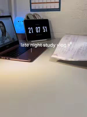 A desk setup with a laptop showing a person on the screen, a clock displaying the time 21:07:51, a blue cartoon-like light, and a piece of paper. The scene is well-lit with warm tones.