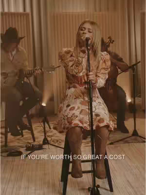 The image captures a musical performance on stage. A woman, wearing a floral dress and boots, is seated and singing into a microphone. A guitarist is seated on the left wearing a cowboy hat. Another musician is in the background playing a stringed instrument. The backdrop is a warm-toned indoor setting.