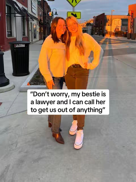 Two women stand on a city street, facing the camera, near a storefront and a trash can. One woman is blonde and wearing glasses, and the other has dark hair. A text overlay appears at the bottom.
