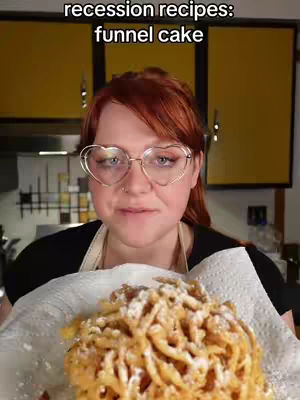 A woman with red hair and heart-shaped glasses holds up a plate containing a funnel cake covered in powdered sugar. Behind her are yellow cabinets and a glimpse of a kitchen setting. The woman is facing the camera, looking directly at the viewer.