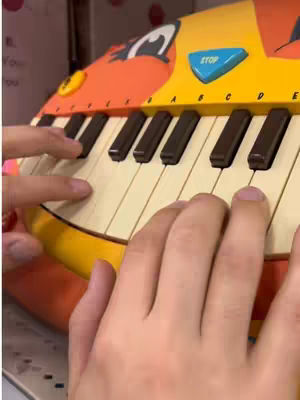 A close-up shot shows two hands playing a colorful toy piano. The piano is primarily yellow, orange, and red, with black and white keys. The hands are positioned over the keys, with fingers pressing down on the keys.