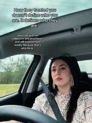 A woman is driving in a car, seen from the front passenger seat. She has long dark hair, is wearing a floral print top and large earrings, and is looking forward with a thoughtful expression. Text overlays appear on the car's windshield.