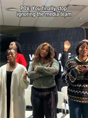 A group of five women, with different hairstyles, standing in what appears to be a church building. The woman in the center has her arms crossed and is looking directly at the camera. The woman on the left has her arms at her side and a white cover up. The woman on the right side is wearing a sweater with a large design. The background appears to be a large sign with white letters.