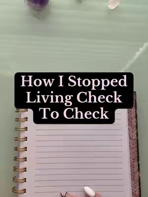An overhead shot shows a partially open spiral notebook on a table. Above the notebook, there's a black banner with white text, reading, "How I Stopped Living Check To Check." The notebook is lined with a hand holding a pen in the lower right corner. The background is a light, solid color with a few scattered objects.