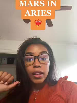 A person with dark hair and glasses is centered in the frame, speaking to the camera. Above them is a stylized graphic with text that reads "MARS IN ARIES"