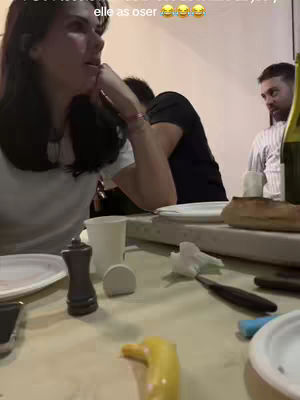 A group of people are gathered around a table, engaged in conversation. The main focus is on a woman on the left side of the table, who appears to be listening or responding with a thoughtful expression. The table is set with plates, cups, and food items, suggesting a meal or social gathering.