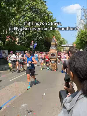 A crowd of marathon runners, with a female runner in the foreground wearing a blue top and black shorts. Next to her is a person wearing a large Big Ben costume.  Behind them are more runners and spectators, with trees and a tall building in the background.