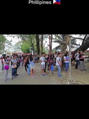 A group of people lined up. The image is in a vertical format with a black background. In the top center, there's text in white that reads "Phillipines" followed by the flag of the Philippines. The main focus is a crowd of mostly young people standing outdoors on a sidewalk or pathway. The people are of diverse complexions and varied attire, appearing to be waiting or lined up. Trees line the background, and the lighting suggests daytime. The image's overall composition is straightforward, centered on the crowd.