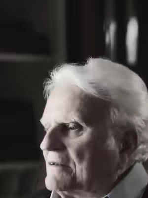 A close-up shot of an elderly man with white hair, looking slightly off-center to the left. He is wearing a dark suit and a white dress shirt. The background is a soft, dark space with indistinct shapes, suggesting a room interior. The image has decent lighting, highlighting the man's face and features.
