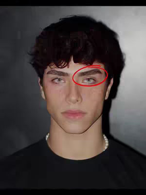 A close-up shot of a young man's face. He has dark hair styled with loose curls, and wears a black t-shirt. The background is a simple, dark gray. His facial features are well-defined, and there's a focus on his eyebrows and eyes, highlighted by a red circle. A pearl necklace is visible at his neck.