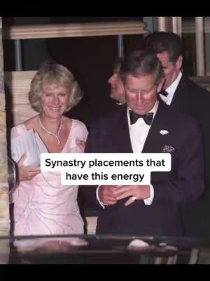 The image showcases a formal event with two main figures: Camilla Parker Bowles and King Charles. Camilla, on the left, is wearing a light pink, embellished dress and smiling, seemingly engaged in conversation. King Charles, to her right, is dressed in a black tuxedo with a bow tie, appearing to be looking down. Other people are visible in the background in formal attire. The setting has a classic and slightly aged feel, likely an indoor event with hints of architecture forming the backdrop