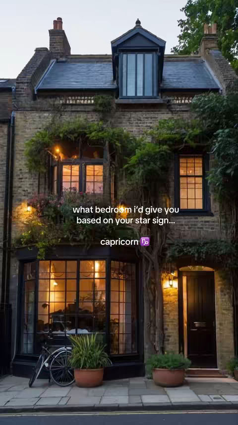 A photograph depicts a three-story brick building, likely a house, with a dark slate roof. The facade is adorned with climbing plants, and the windows have lights emanating from inside. Text overlays on the image read, 'what bedroom i'd give you based on your star sign...' and 'capricorn'. The overall composition creates a cozy, inviting atmosphere with warm lighting.