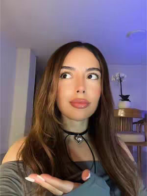 A woman with long brown hair looks to the side, with one hand extended towards the camera. She's wearing a black choker and a gray top.