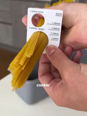 A person's hand holds a white plastic tool with the measurements for pasta. The tool shows holes where pasta is placed. The pasta and tool are held in front of a blurred background showing another container.