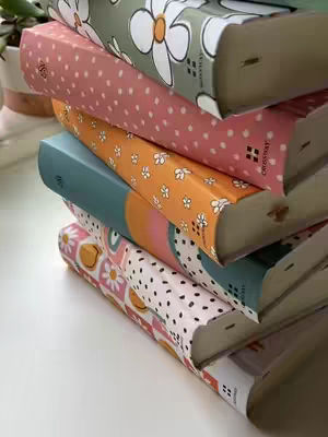 A close-up shot of a stack of ESV journaling Bibles, with patterned covers in various colors and designs. The books are arranged in a diagonal stack, showing the top edges and spines. The covers feature floral and polka dot patterns, with colors including green, pink, orange, and blue. The lighting appears natural, highlighting textures and colors.