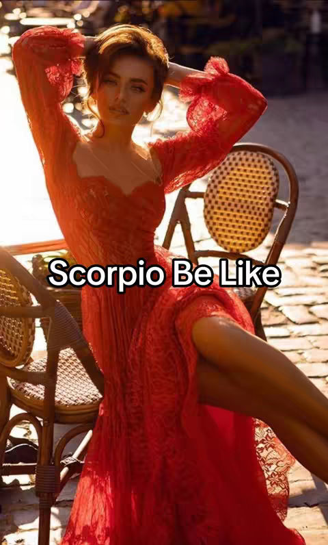 A woman with fair skin and long brown hair is the central figure in the image. She is wearing a long, vibrant red dress, standing outdoors. Her arms are raised above her head, with her hands touching her hair, suggesting a pose of relaxation or emphasis. The background features a partially visible patio setting, with wooden chairs and soft, natural light, creating a warm and inviting atmosphere. Overlayed text at the front spells out 'Scorpio Be Like'.