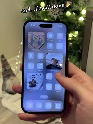 A person's hand holds a smartphone displaying a custom home screen with various app icons, a winter-themed calendar widget, and a Christmas tree in the background. The overall aesthetic is cool-toned and organized.
