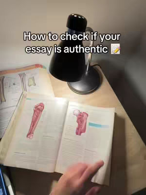 The image shows a desk with an open anatomy book lit by a desk lamp. The hand in the image is turning the page, with the lamp creating soft shadows. The text overlay reads: How to check if your essay is authentic.