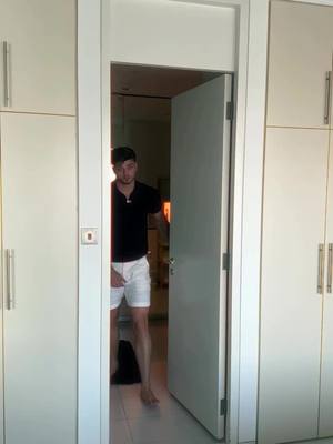 A man is partially visible in an open white door frame, framed by white cabinets. The man, wearing a black t-shirt and white shorts, stands barefoot holding the door open. The background is slightly out of focus, suggesting the image was taken indoors.