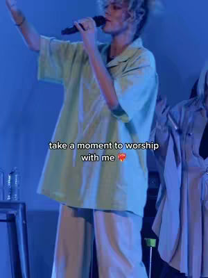 A person with short, light-colored hair is on a stage, holding a microphone and singing. They are wearing a light green shirt and light pants. Another person is standing to their right. The image is lit with cool-toned stage lighting, predominantly blue.