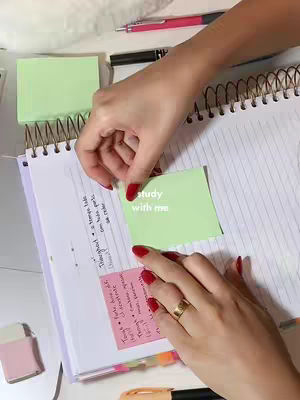 The image shows a person's hands, with red nail polish, interacting with a notebook and sticky notes on a desk. A green sticky note reads 'study with me'. There are pens and a notepad on the desk.