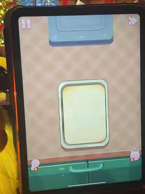 A screenshot of a mobile game in landscape mode. The top of the screen shows a light blue oven. Below the oven is a light tan wall. In the center is a rectangular tray with a yellow surface. Beneath the tray is a green kitchen cabinet. Two pink icons are in the top left and two red icons with clocks are in the bottom right. The game is being displayed on a tablet.