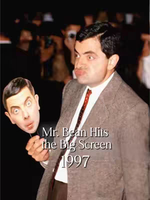 The cover image features Rowan Atkinson, in character as Mr. Bean, posing with a Mr. Bean mask. He is wearing a brown suit and a red tie, with a surprised expression on his face and his lips puckered. Text overlays the image in the style and read "Mr. Bean Hits the Big Screen 1997". The background is blurred, likely from a red carpet or event.