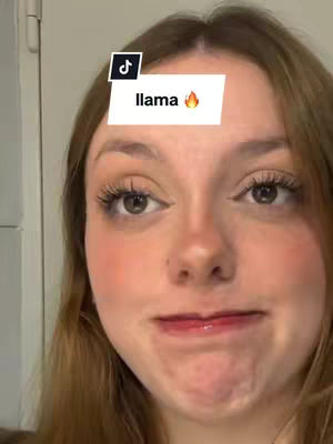 A close-up shot of a young woman's face. She has fair skin, long eyelashes, and light brown hair. A superimposed graphic on her forehead reads "llama" with an emoji of a flame. She has a slightly pursed expression.