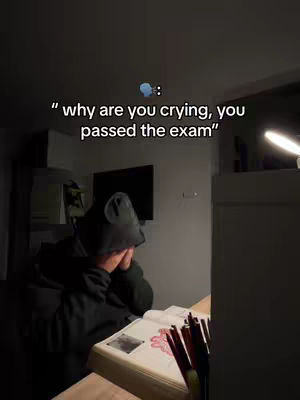 A person, possibly a student, is sitting at a desk in a dimly lit room, with their head in their hands, looking upset. A book sits open on the desk, and there are writing utensils visible. A desk lamp provides light, creating a contrast between the lit area and the shadows.