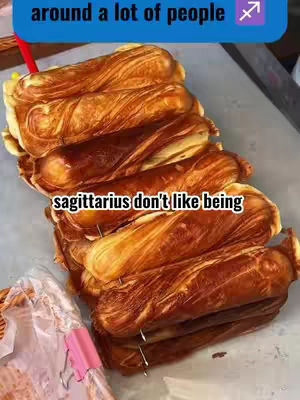 A top-down shot of a stack of golden-brown pastries, possibly croissants or similar, with a red straw and a pink clip visible in the background. Text overlays in blue and white state 'around a lot of people' and 'sagittarius don't like being'.