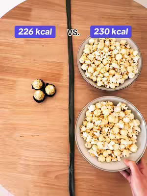 An overhead shot shows a wooden table divided by a long, dark strip. On the left are three chocolate candies with gold wrappers. On the right are two bowls of popcorn, situated below each other, with a hand partially visible at the bottom, holding one. The top left corner features a blue rectangular text overlay with white text, and the top right corner has a similar overlay.