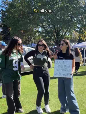 Three young women stand outdoors. The woman on the left is wearing a green and white sports jersey with the number 9. The woman in the center is wearing a black top and black pants and has her arms crossed. The woman on the right holding a sign that says "SAY 5 NICE THINGS ABOUT YOUR OPPONENT FOR 6TH (???)"