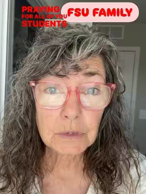 A close-up shot of a woman with curly gray hair and pink-framed glasses. Text overlays state 'Praying for all of you students' and 'FSU FAMILY'. The background is a blurred indoor setting with a hint of a door.