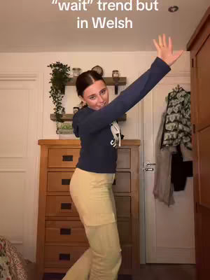 A woman in a blue hoodie and tan pants stands in a room with a wooden dresser, shelves with decor, and a door. The woman has one arm raised and is positioned as if she is dancing.