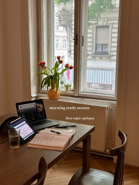 The image showcases a study space with a view of a city through a window. On a wooden table, there's a laptop displaying a music app, and a tablet displaying notes or a document, accompanied by a glass of water, books, and pens. A vase with red tulips adds a pop of color. The room is brightly lit and appears to be a cozy study zone.