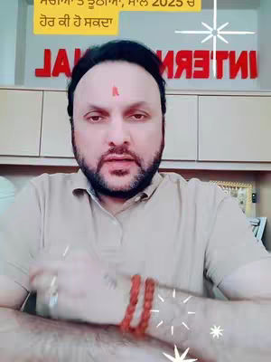 A man with a red mark on his forehead is in the center, looking directly at the camera. The background features a logo and decor, with some text in the top left corner. The man is wearing a light brown shirt and has a beard and mustache. His arm with a bracelet is visible, with some animated star effects are at the bottom and top right.