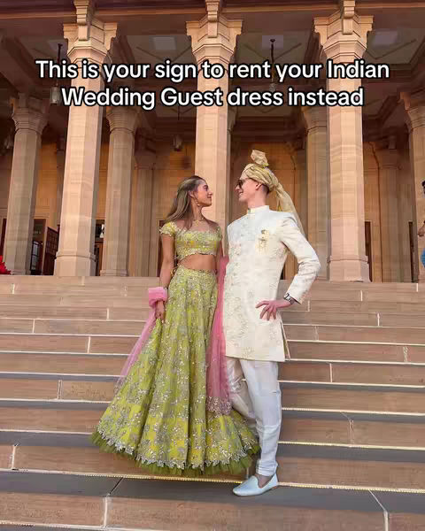 A couple stands on grand steps in front of a large building. The woman is dressed in a green and gold lehenga, looking towards the man. The man wears a cream-colored suit with a turban. Text overlay reads 'This is your sign to rent your Indian Wedding Guest dress instead'