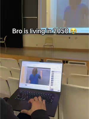 A person is holding a laptop in what seems to be an auditorium. The screen of the laptop displays a video of a person in a blue shirt on a stage. Above the laptop, text overlays read, "Bro is living in 2050". The background includes rows of white chairs and a stage.