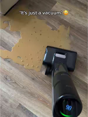 The image shows a black vacuum cleaner pointed towards a large spill of a brown liquid on a light-colored wooden floor. Above, in a white text box, are the words: 'It's just a vacuum'.