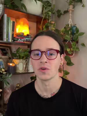 A person with glasses looks directly at the camera, with a background of a shelf with plants, books, and a light.
