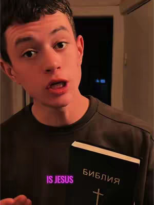 A young man with short dark hair looks directly at the camera, his mouth slightly open as if speaking. The book he holds reads, "БИБЛИЯ" on the cover. In the image's lower left, a text overlay reads "IS JESUS". The background is dimly lit.